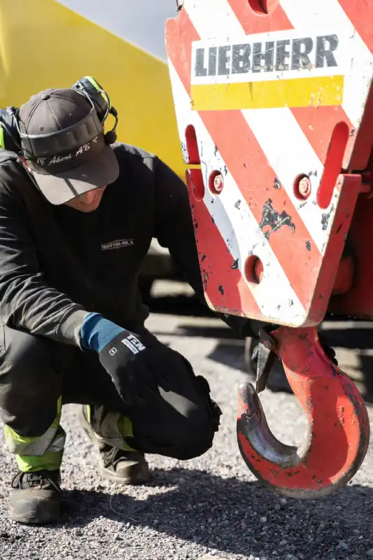 A certified machinery appraiser wearing Personal Protective Equipment (PPE), including ear protection, work gloves, and high-visibility trousers, crouches on a gravel surface to conduct an on-site inspection of a large red Liebherr crane hook. The technician is focused on the crane's pulley block, which features red and white safety stripes, performing a detailed condition assessment for a heavy equipment appraisal.
