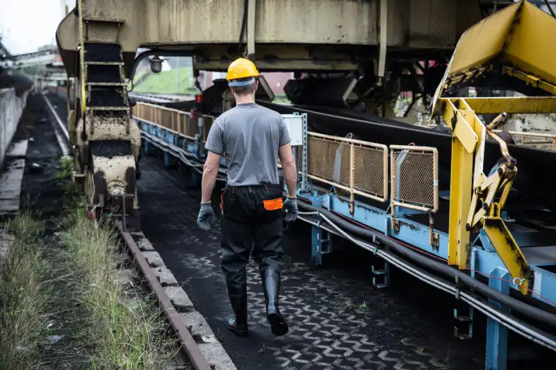 An industrial appraiser wearing a yellow hard hat and PPE safety gear walks alongside a large-scale conveyor belt system and material handling machinery. The scene shows the technician performing an on-site condition assessment of heavy industrial infrastructure, with the conveyor tracks and surrounding steel structures covered in dark material like coal or ore, typical of a mining or power plant equipment valuation.