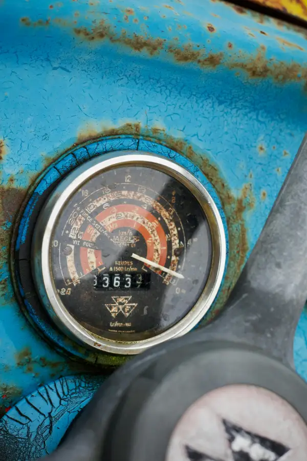 A close-up, top-down photo of a vintage Massey Ferguson tractor's dashboard, focusing on a weathered Jaeger hour meter and tachometer. The gauge, displaying 33,633 hours, is set against cracked and rusted light blue paint, with the Massey Ferguson triple-triangle logo visible at the bottom of the dial. This detail illustrates the critical process of verifying machine hours and usage history during an on-site heavy equipment appraisal.
